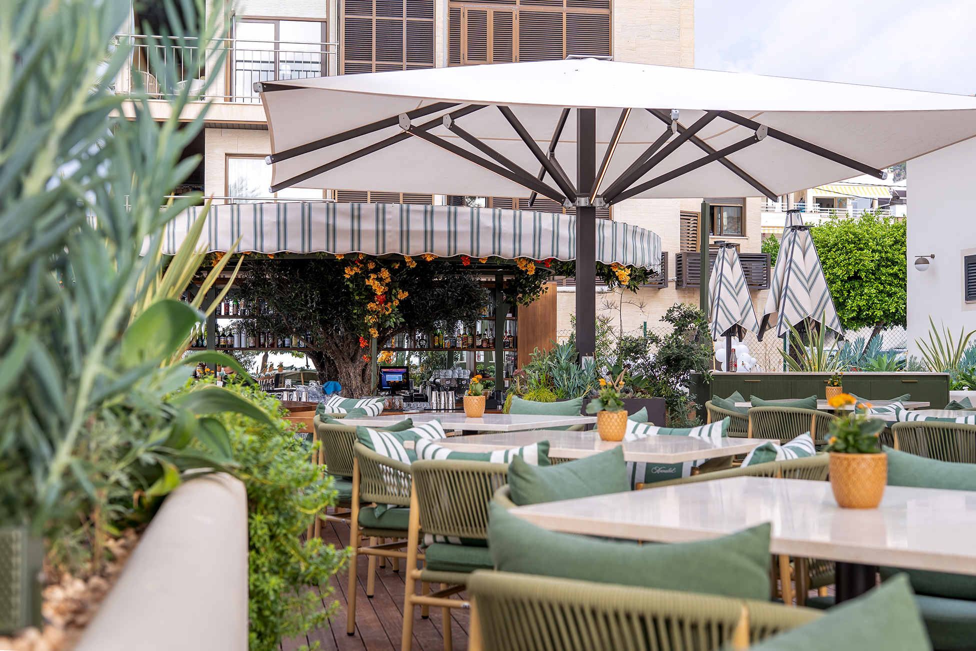 Terraza con mesas y sillas verdes, sombrilla grande y bar con decoración floral bajo toldo rayado.