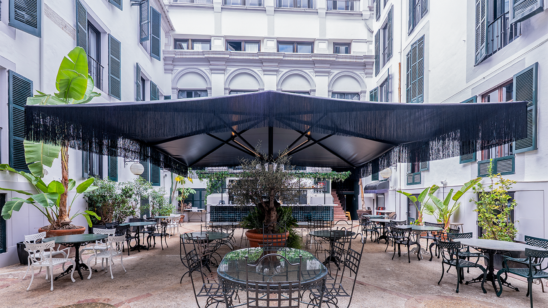 Terraza interior con mesas, sillas de metal, plantas y toldo negro con flecos en edificio.