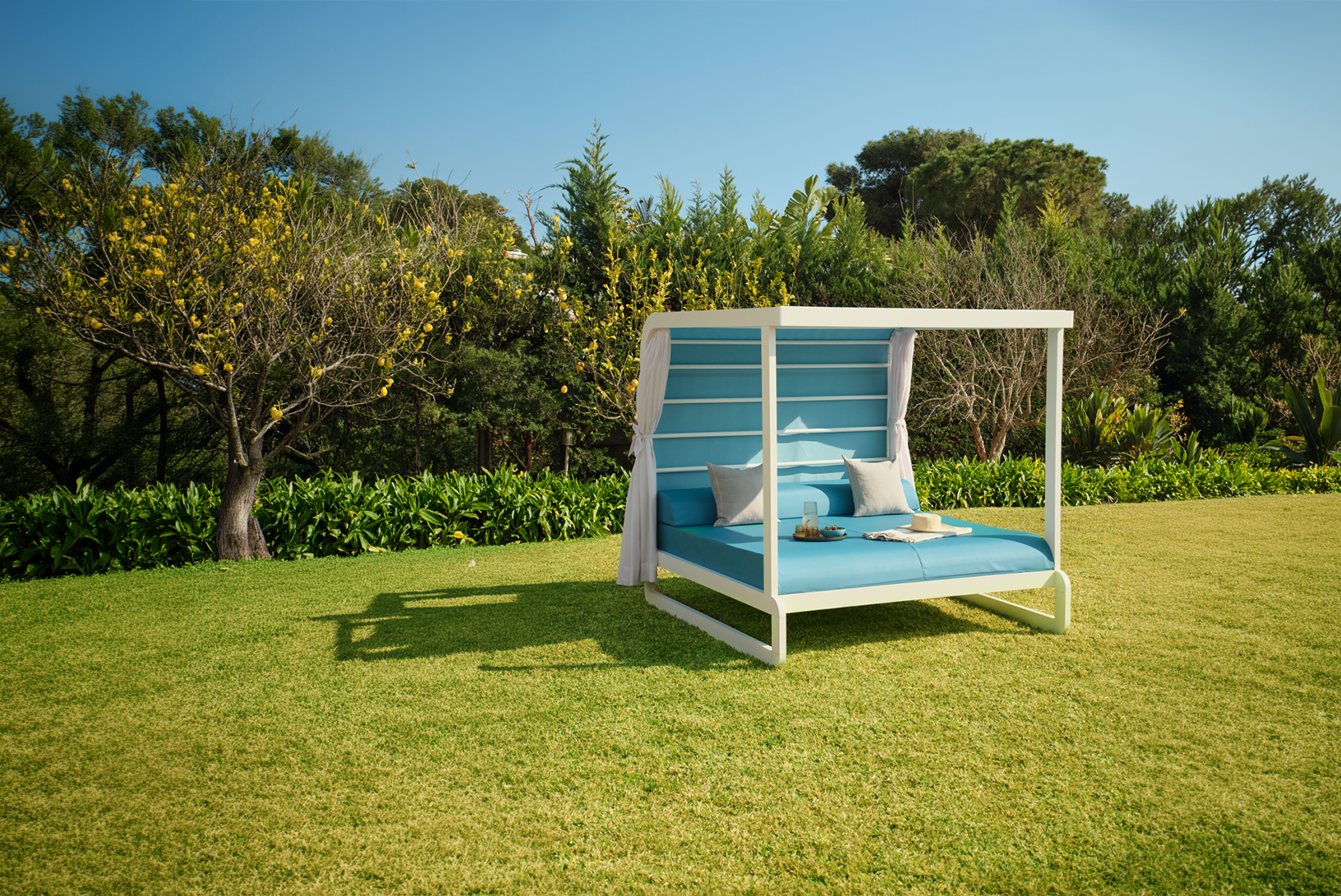 Cama balinesa blanca con acolchado azul, cojines y bandeja en césped con árboles y cielo despejado.