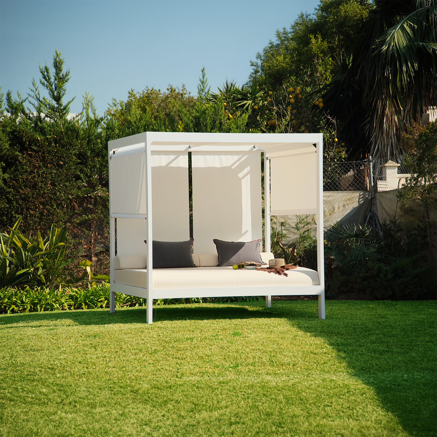 Cama exterior con estructura blanca, cojines grises, sombrero y plato sobre césped verde en jardín soleado.