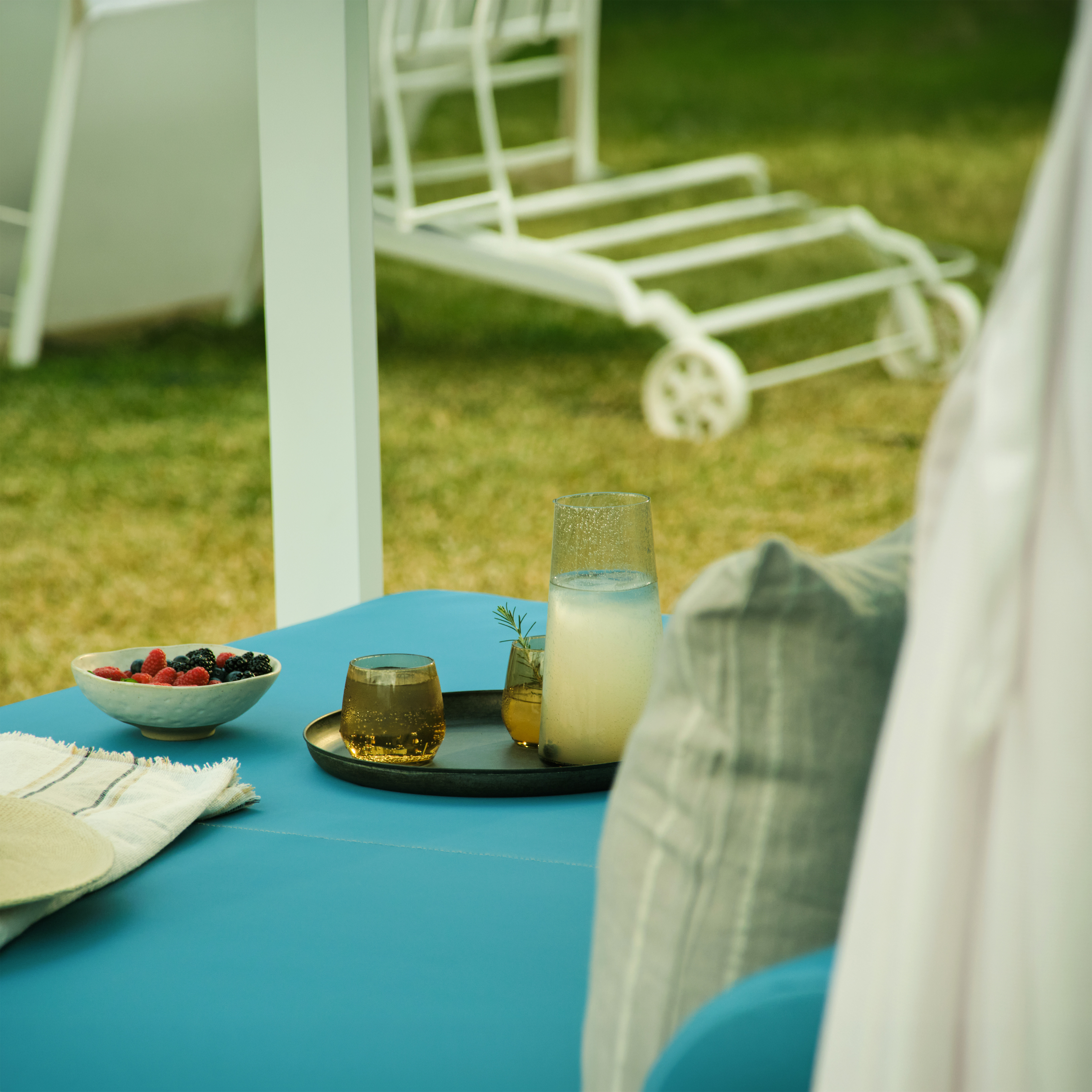 Mesa azul con frutas, jarra y vasos con bebidas en un jardín con tumbona blanca al fondo.