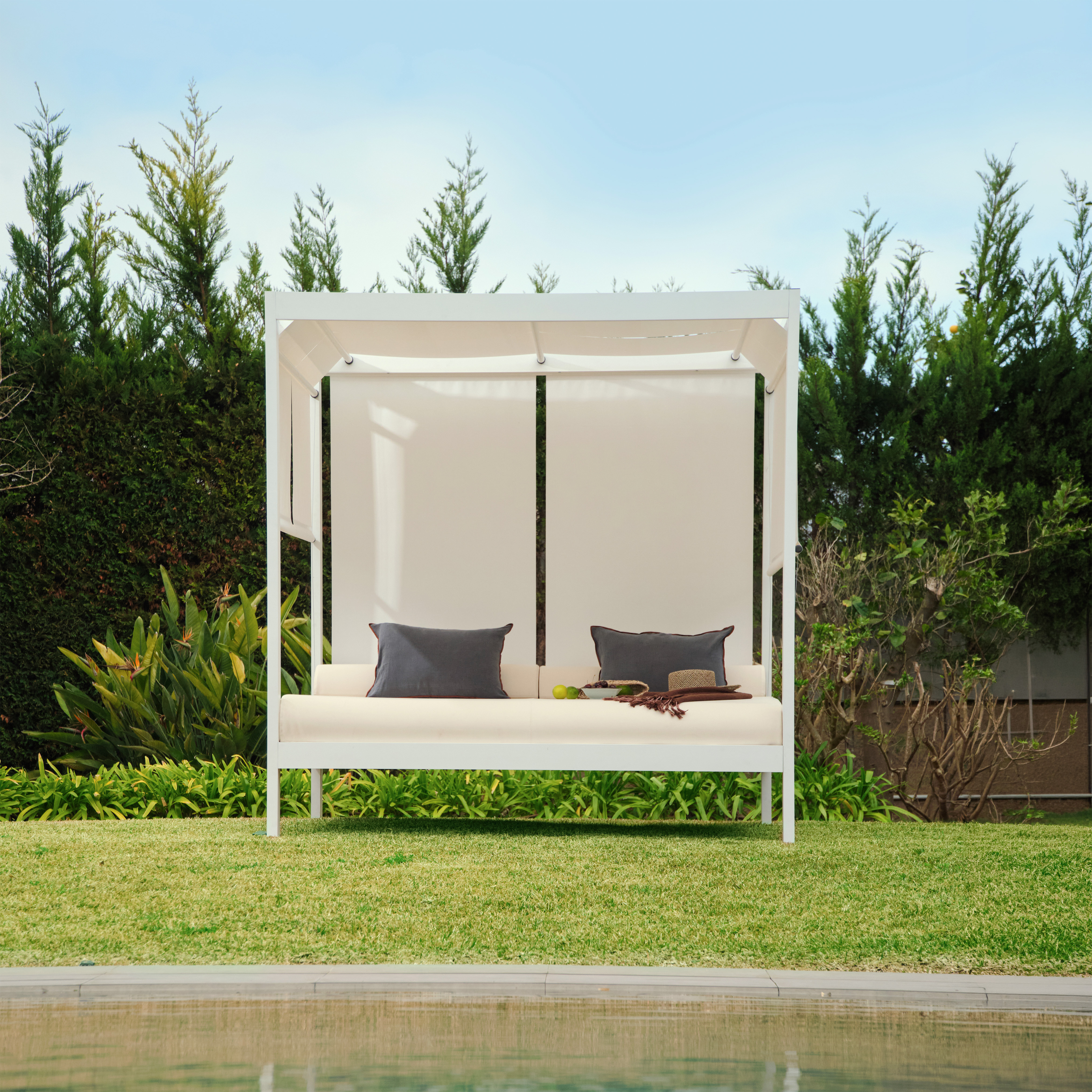 Cama balinesa blanca con cojines junto a una piscina en jardín con césped y arbustos.