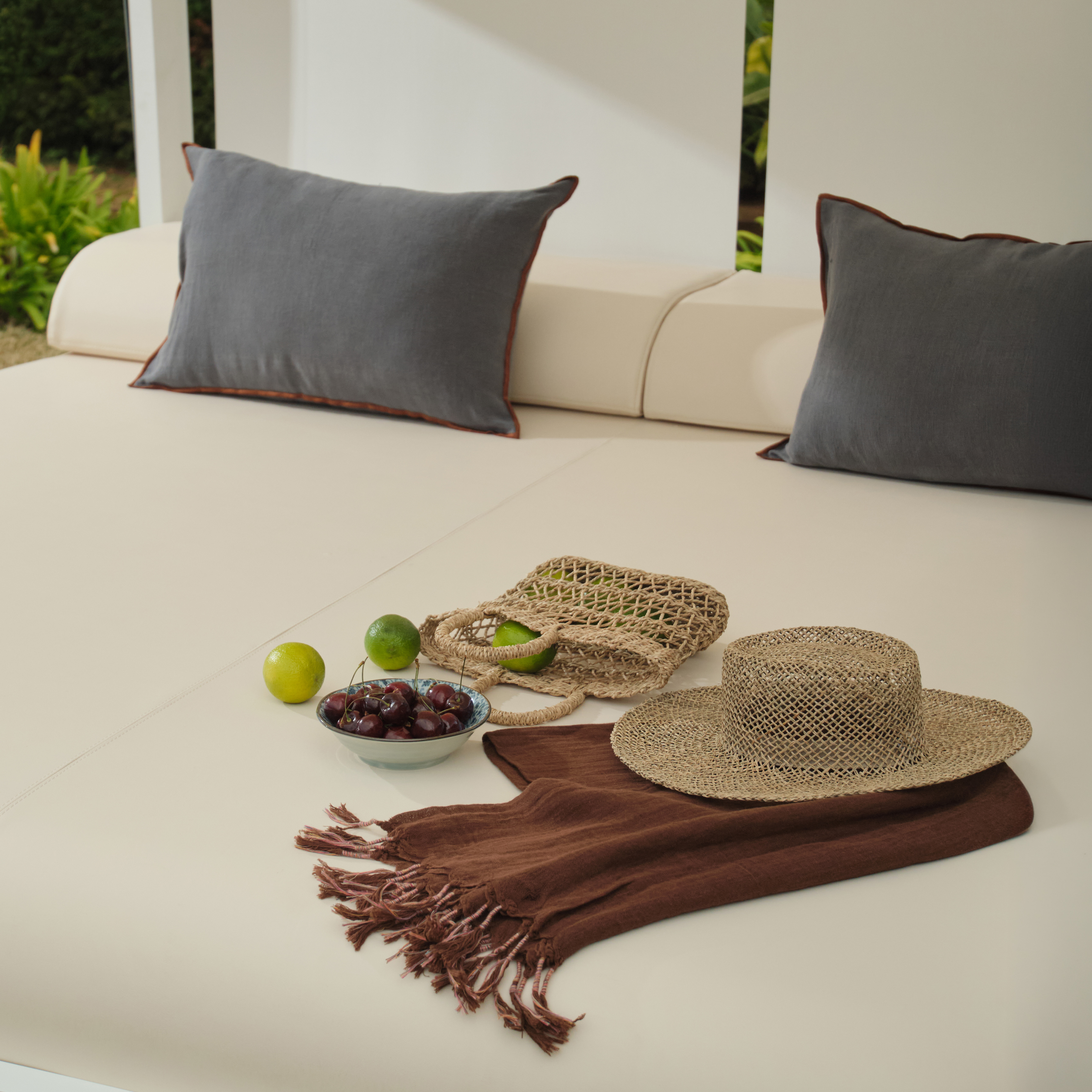 Sombrero de paja, bufanda marrón, bolsa tejida, bowl con cerezas y limones en sofá beige con almohadas.