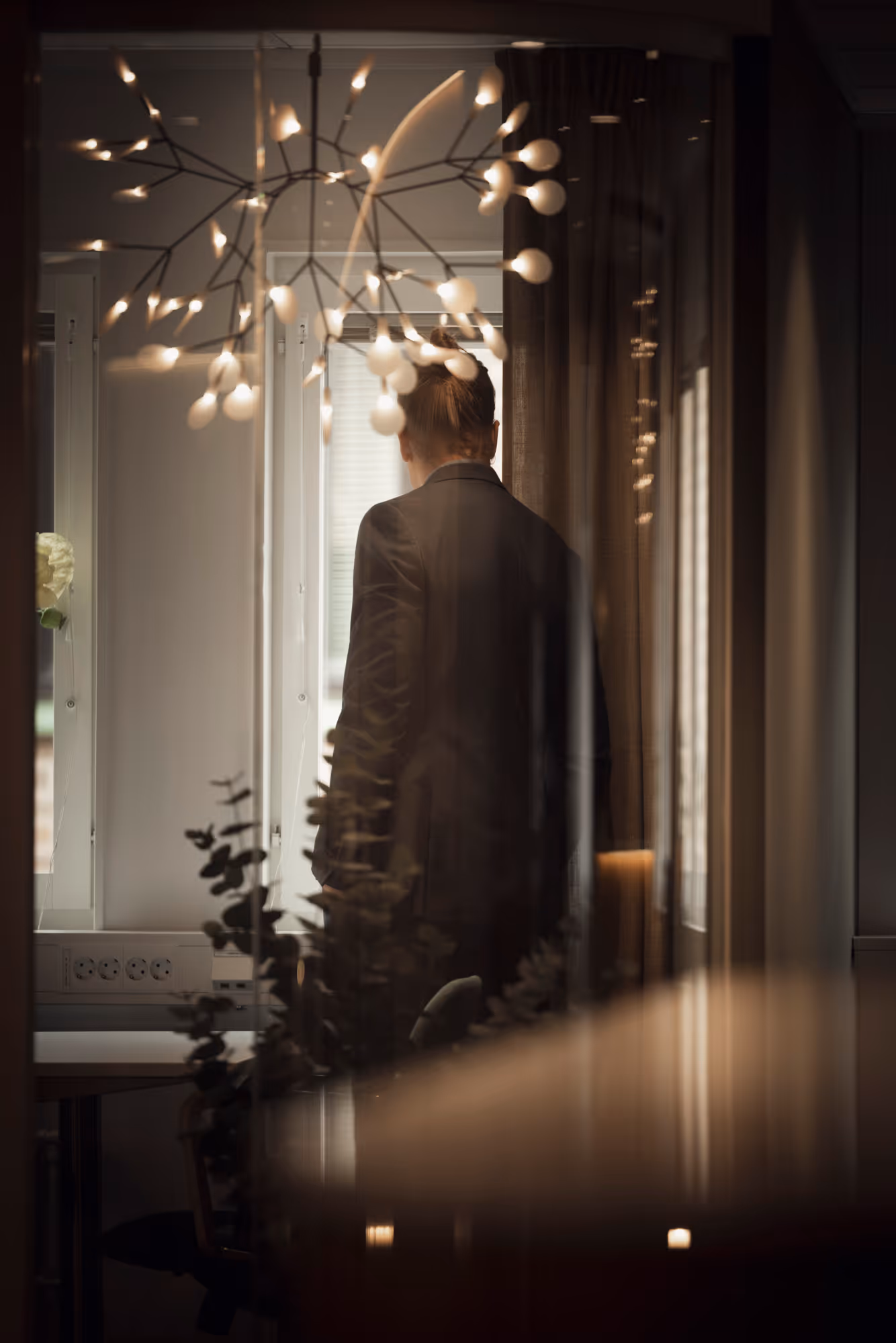 Person i kavaj med uppsatt hår, sedd bakifrån, står vid ett fönster med varmt ljus.