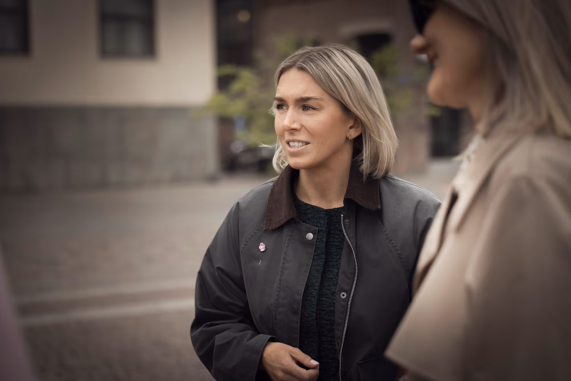 Kvinna med kort blont hår och mörk jacka som står ute och ser åt sidan.