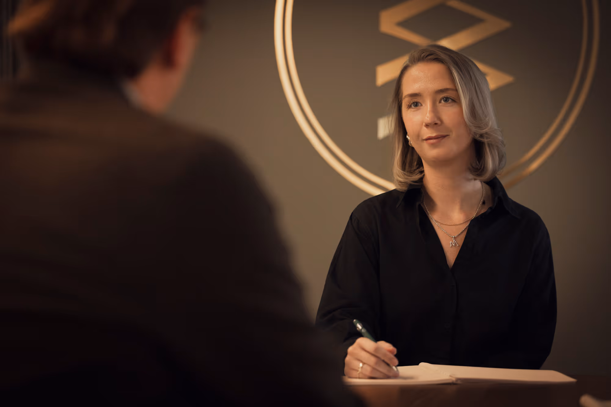 Kvinna i svart skjorta skriver i anteckningsbok under samtal med en person i ett kontor.