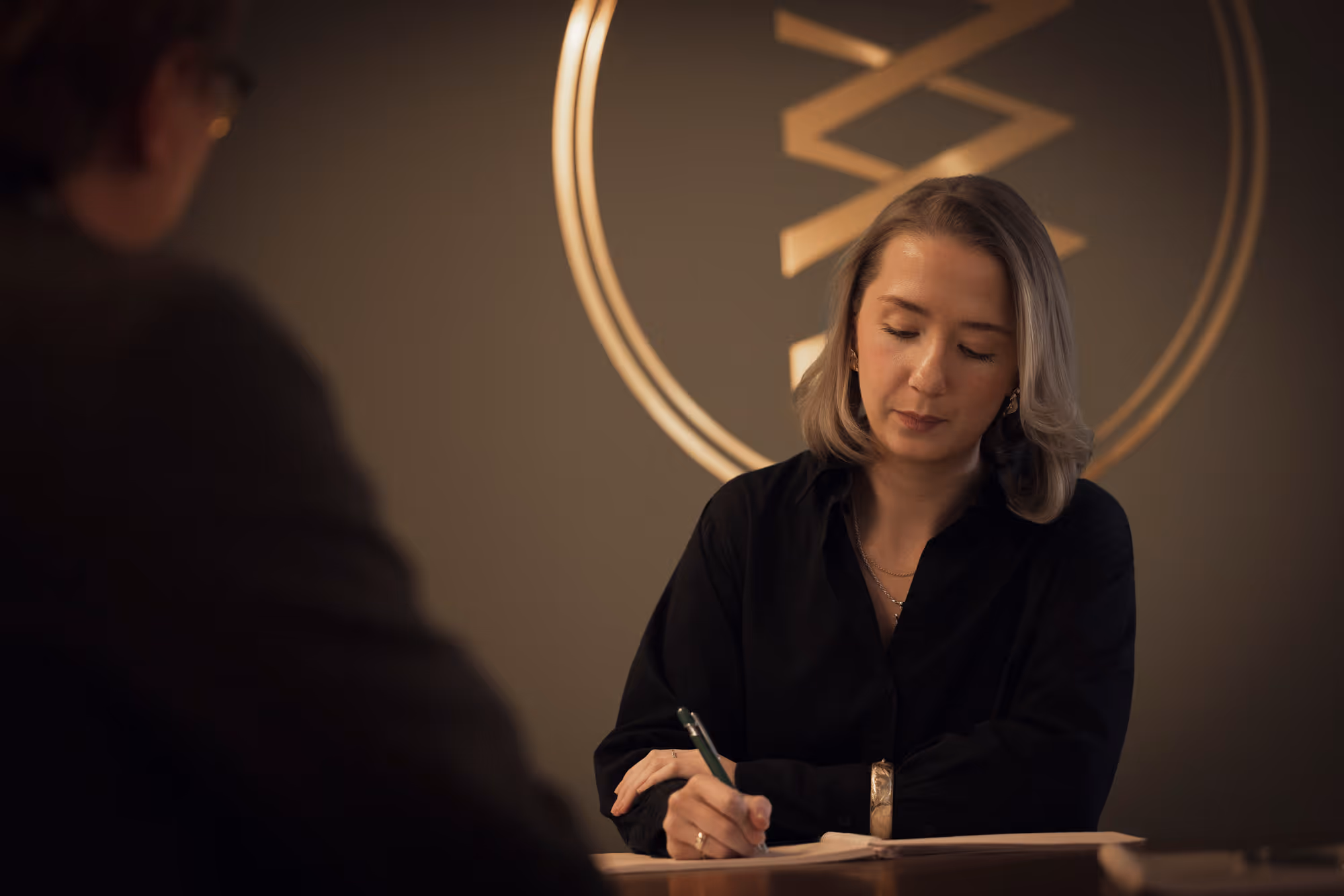 Kvinna i svart skjorta skriver vid ett skrivbord med en person i förgrunden och rund dekoration i bakgrunden.