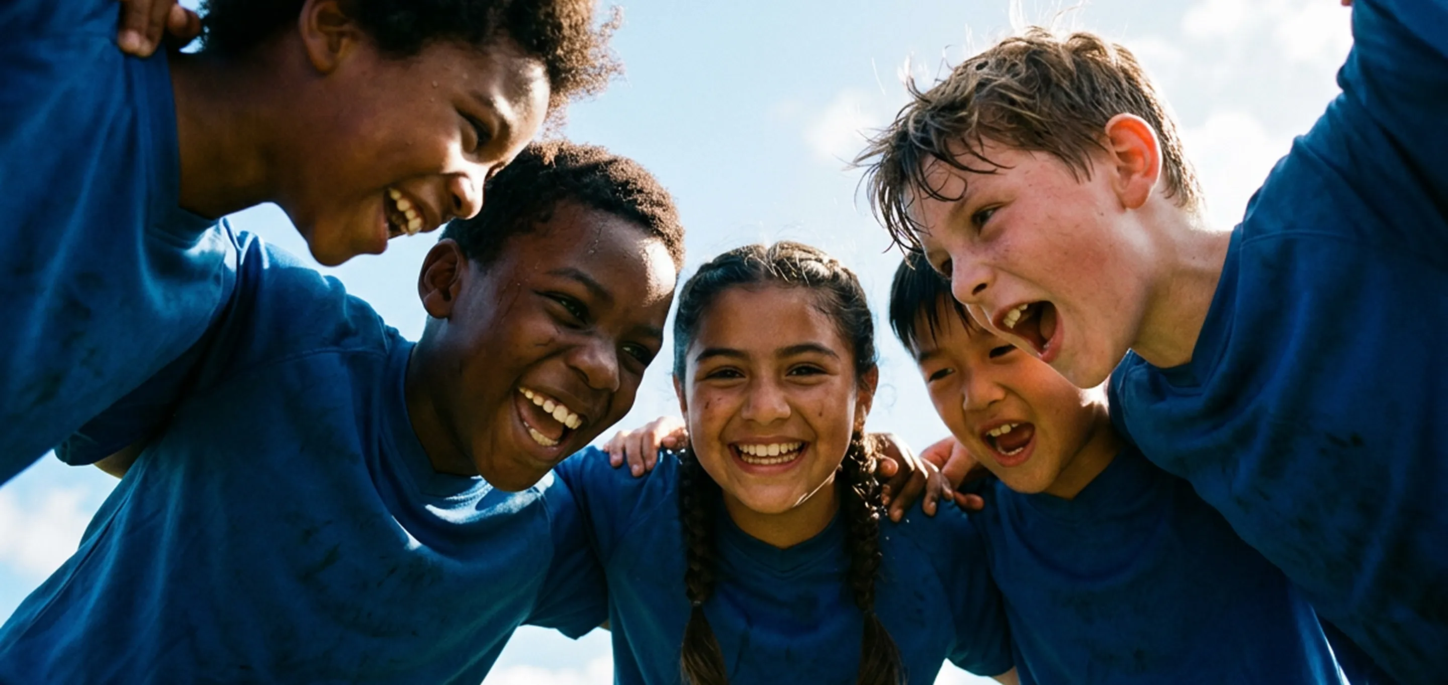 Fünf Kinder in blauen Sportshirts stehen in einer Gruppe mit Armen um die Schultern und lachen gemeinsam.