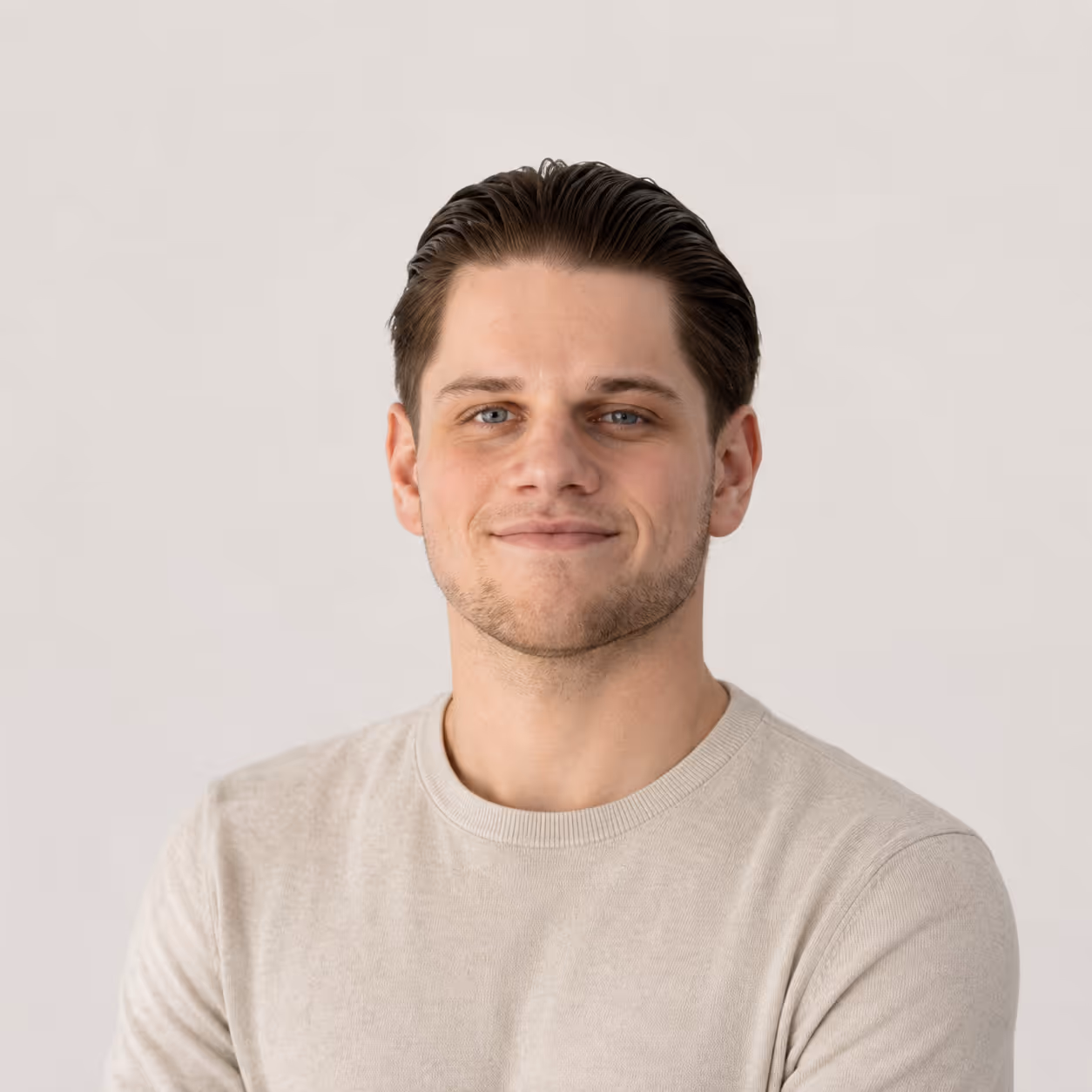 Junger Mann mit dunklem Haar und blauem Blick, lächelnd in cremefarbenem Pullover vor hellem Hintergrund.