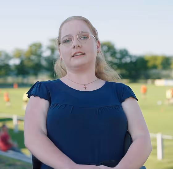 Frau mit Brille und blauem Kleid sitzt draußen vor unscharfem grünen Hintergrund.