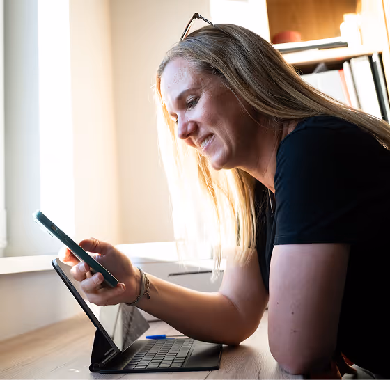 Frau mit langen blonden Haaren lächelt, während sie auf ihr Handy schaut, neben Tablet mit Tastatur.