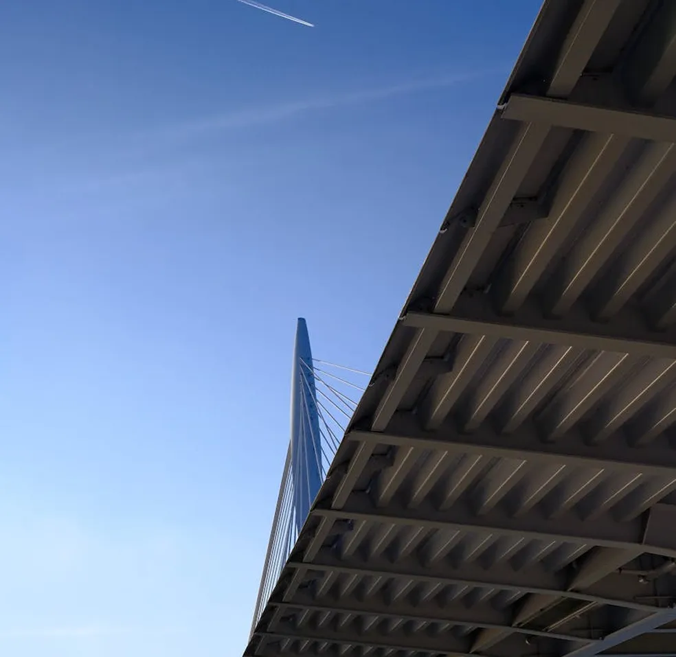 Underside of a modern cable-stayed bridge with a clear blue sky and an airplane contrail above.