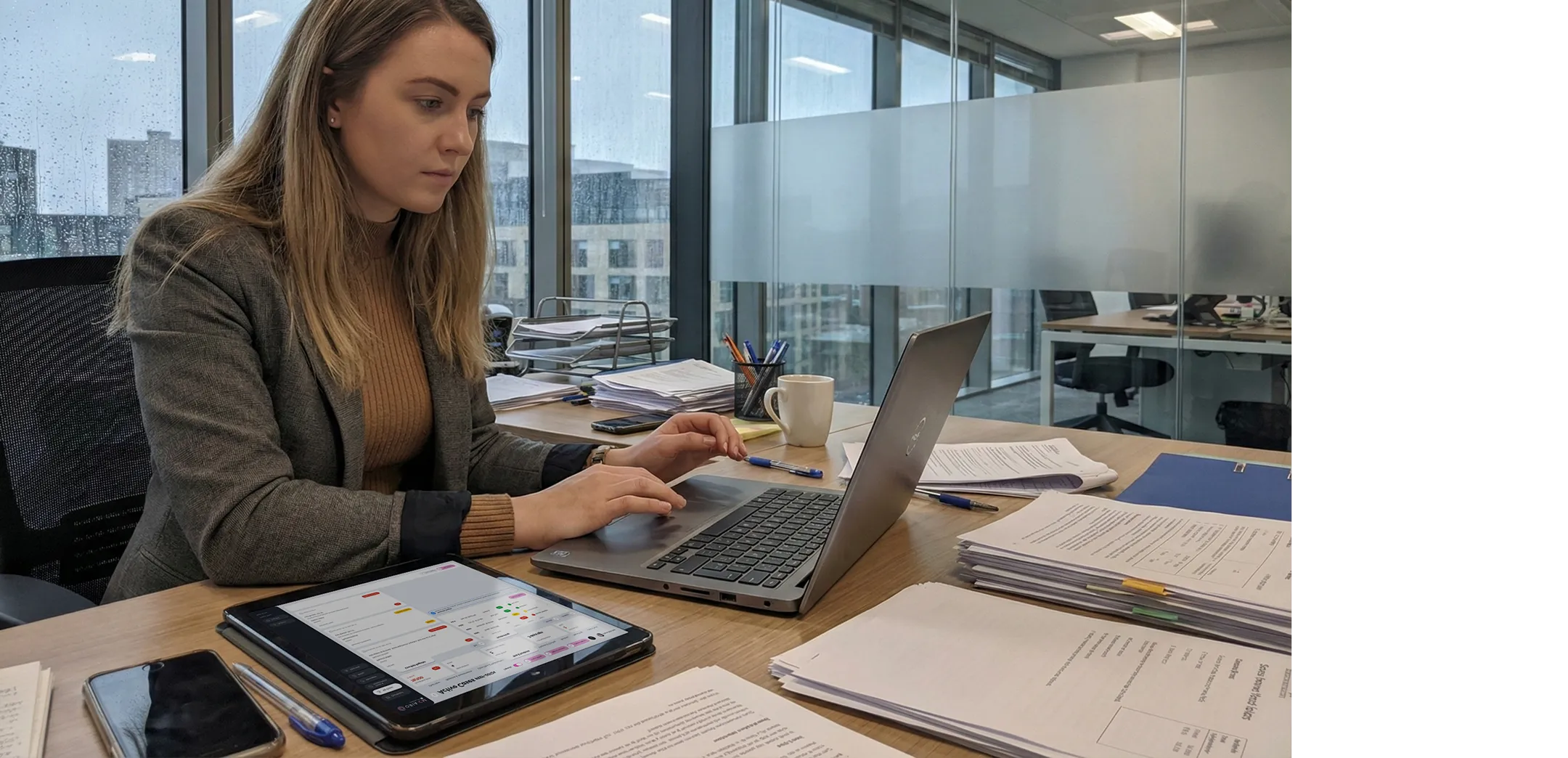 Woman working on a laptop at an office desk cluttered with documents, tablet, pens, and a coffee mug.