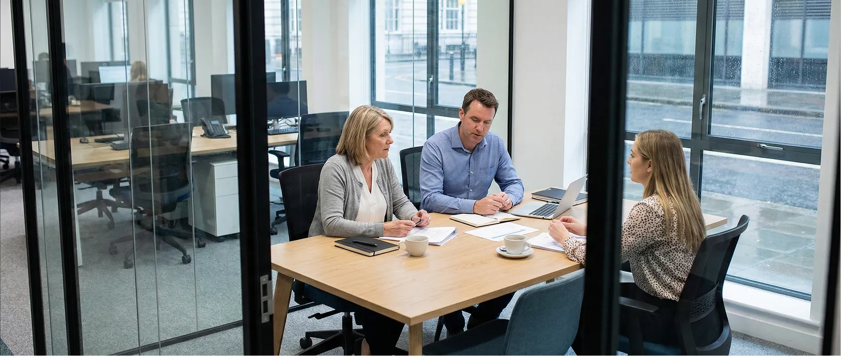 Three business professionals having a meeting around a table with documents and a laptop in a glass-walled office.