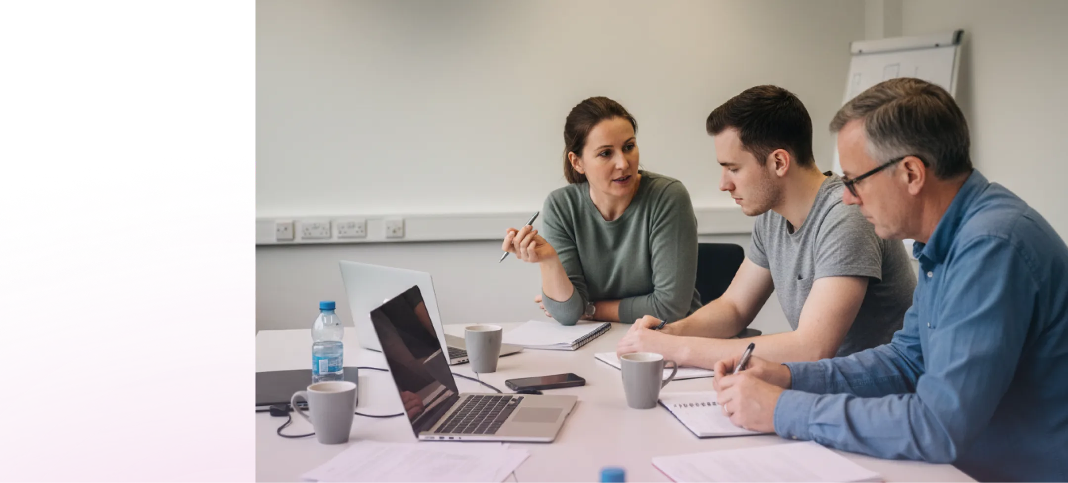 Three colleagues in a meeting room discussing work with laptops, notebooks, and coffee cups on the table.