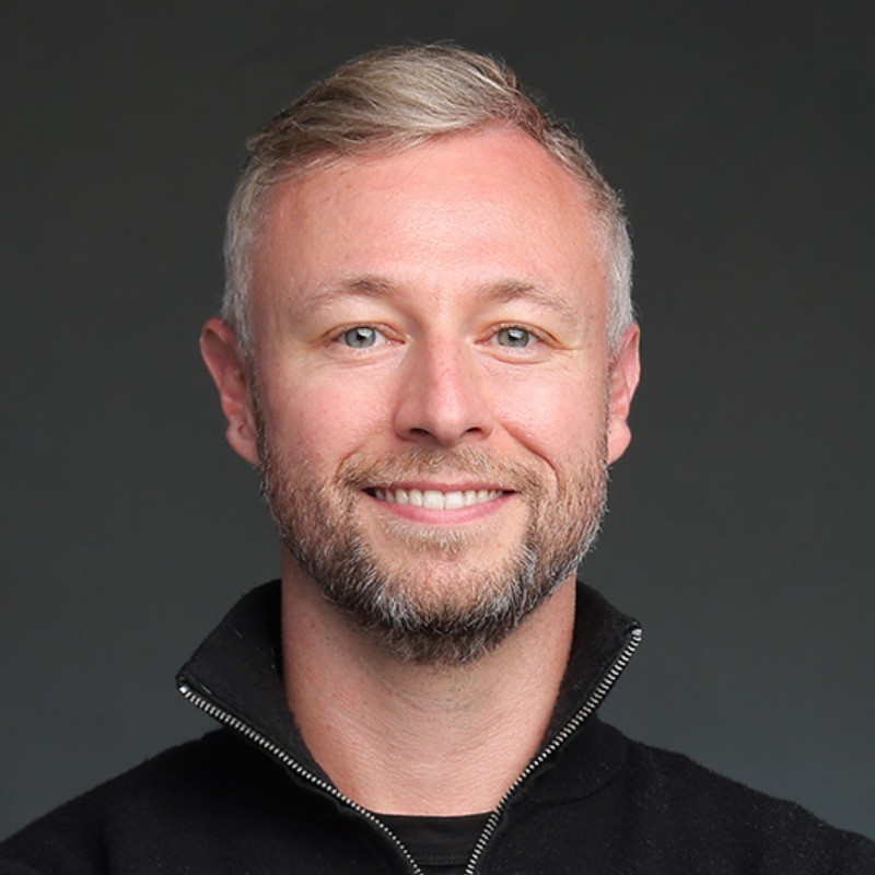 Smiling man with blond hair and beard wearing a black zip-up sweater against a dark background.