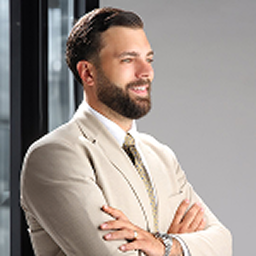 Smiling man with beard and slicked-back hair wearing a beige suit and patterned tie, standing with arms crossed.