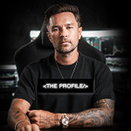 Man with tattoos on arms sitting in front of computer screens, wearing a black T-shirt with text '<THE PROFILE>'.