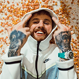Smiling man wearing a white Nike hoodie and a watch, showing middle fingers on both hands with tattooed arms.
