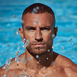 Close-up of a muscular man with short hair emerging from water with droplets splashing around him against a blue background.