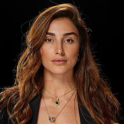 Portrait of a woman with long wavy brown hair wearing two gold necklaces with pendants and a black top against a black background.