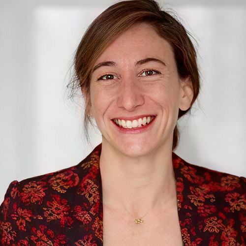 Smiling woman with brown hair wearing a red patterned blazer and a small gold necklace.