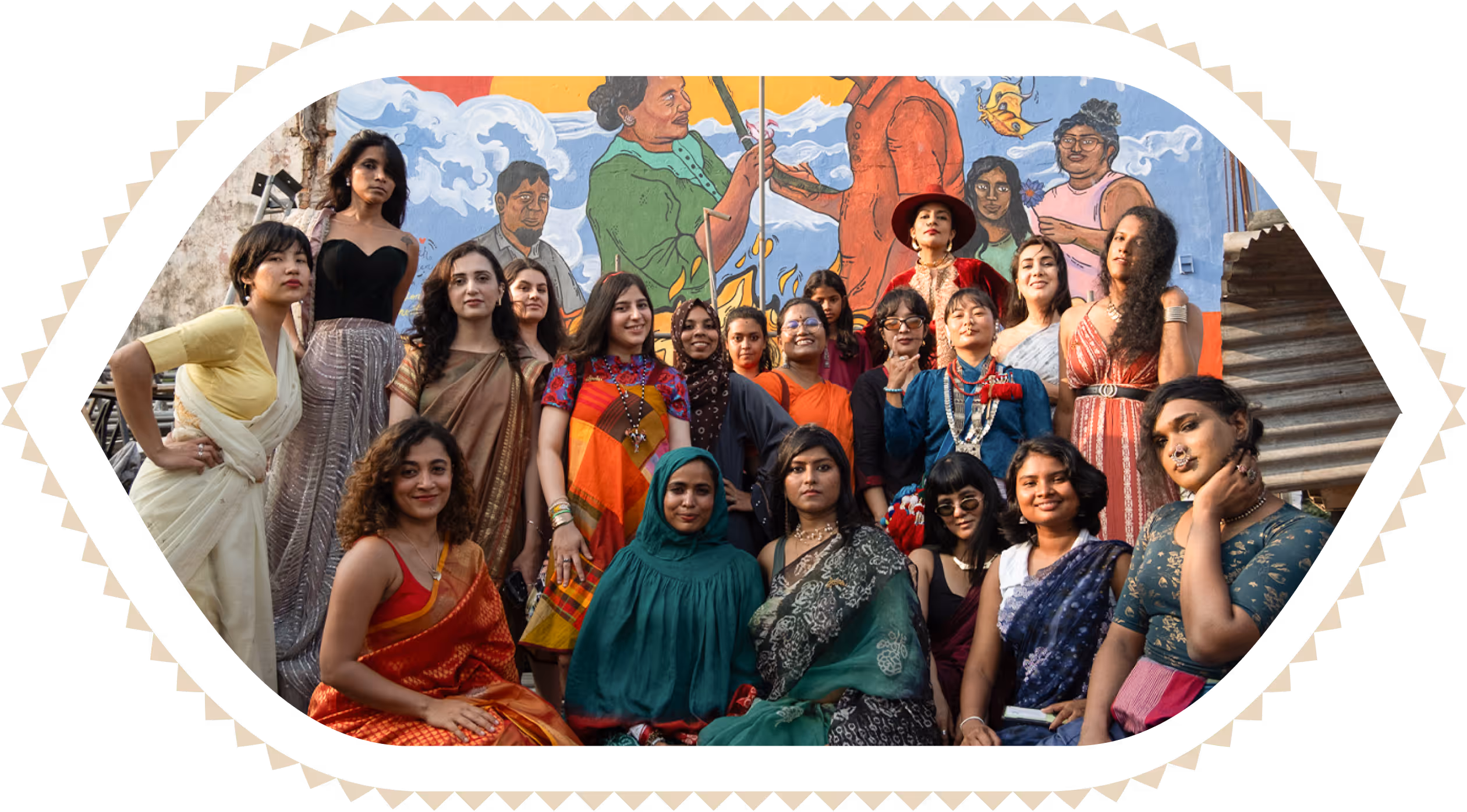 Group of diverse women and gender-diverse people posing in colorful traditional and modern clothes in front of a mural.