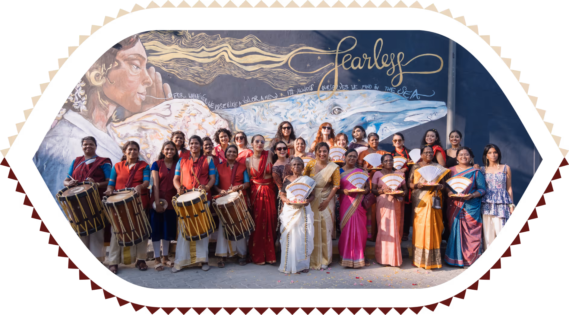 Group of women in colorful traditional attire posing with drums and fans in front of a mural with the word Fearless.