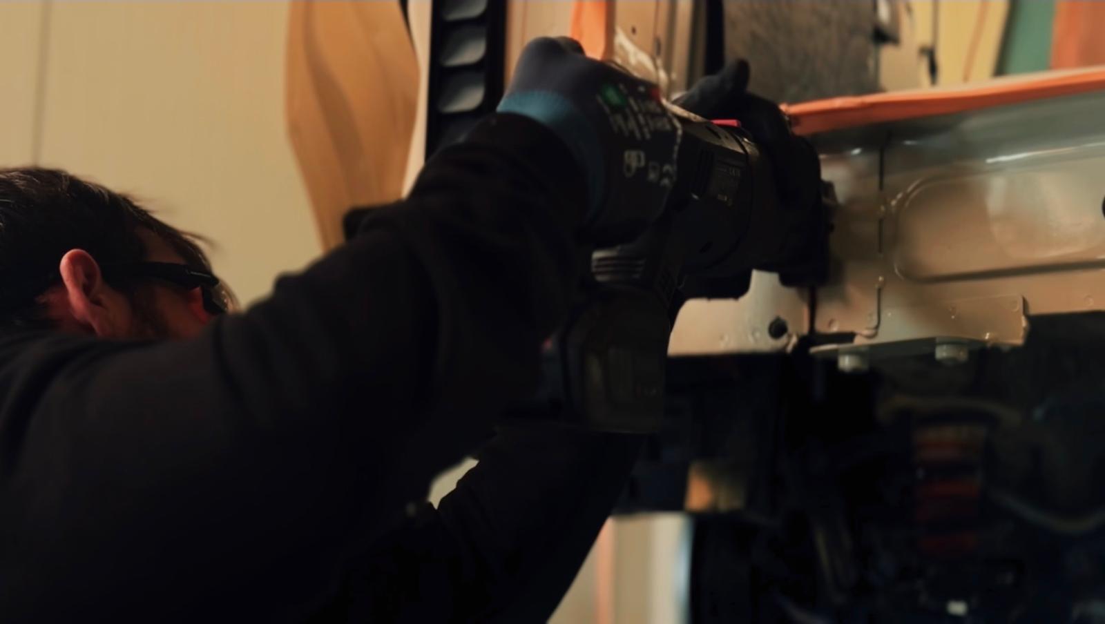 Person wearing gloves using a cordless drill on a light-colored metal surface.