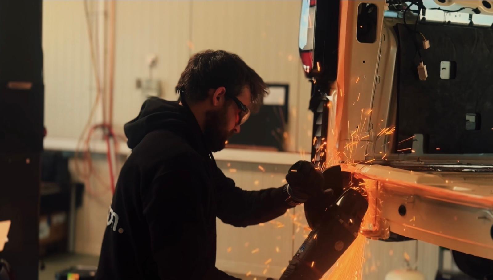 Man wearing safety glasses and gloves using a power grinder to cut metal on a vehicle in a workshop with sparks flying.