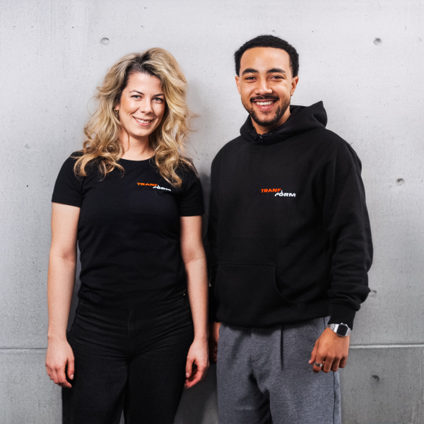 Smiling woman and man standing side by side against a gray concrete wall, wearing black shirts with the word 'TRANSFORM' printed on them.