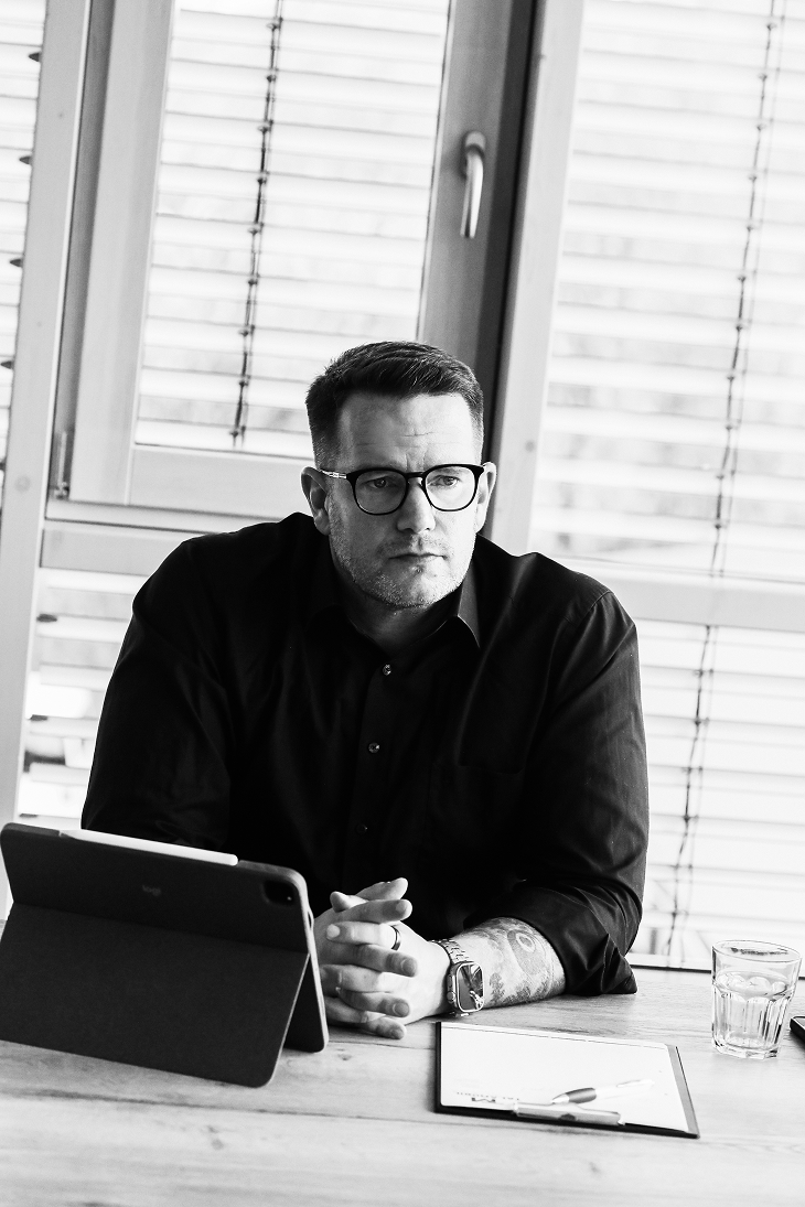 Man with glasses and a tattoo sitting at a desk with a tablet, clipboard, and glass of water in front of a window with blinds.