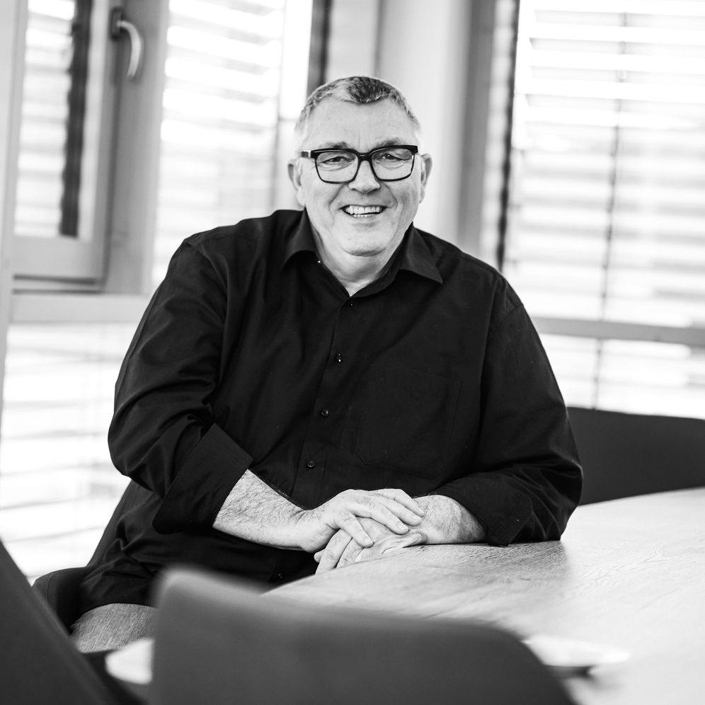Smiling middle-aged man wearing glasses and a black shirt sitting at a wooden table in a room with window blinds.