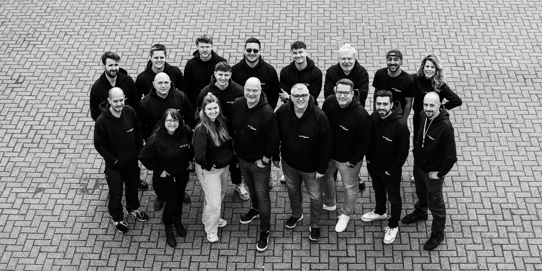 Group of 18 people standing on a paved surface in a staggered formation, wearing matching black hoodies.
