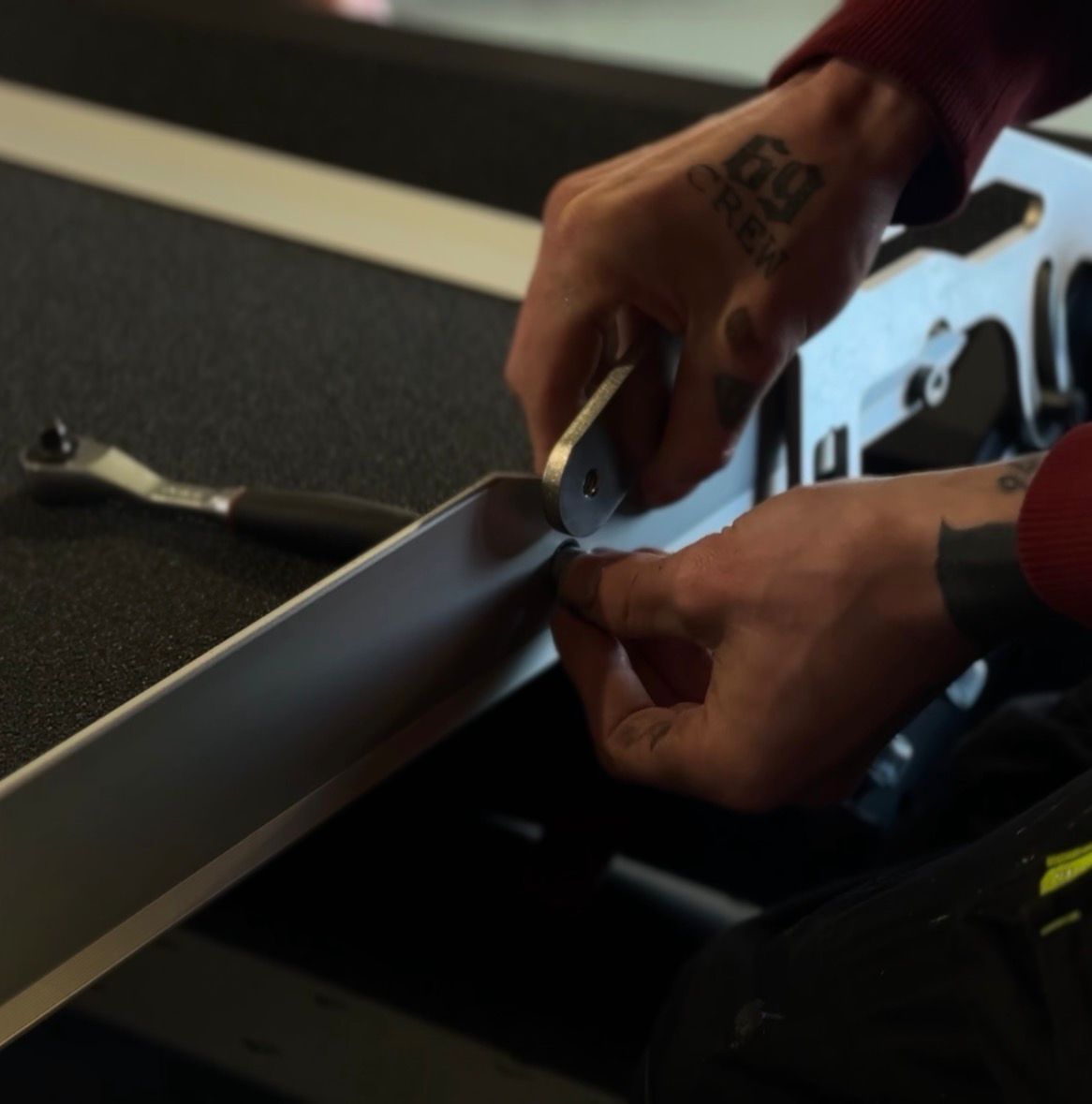 Hands with tattoos using a tool to tighten a bolt on a metallic frame.
