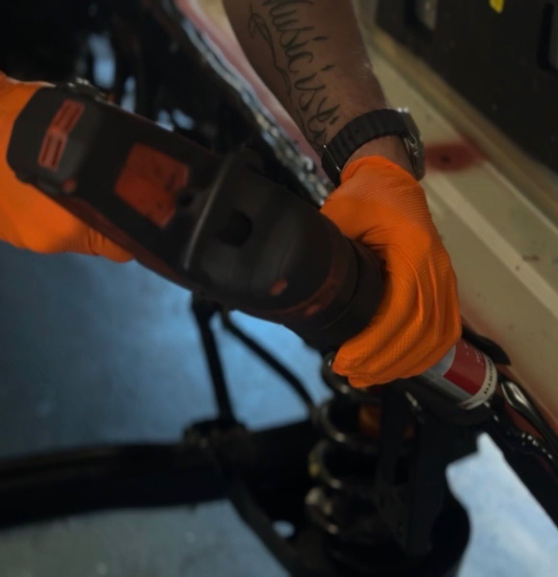 Person wearing orange gloves using a power tool to work on a vehicle suspension.