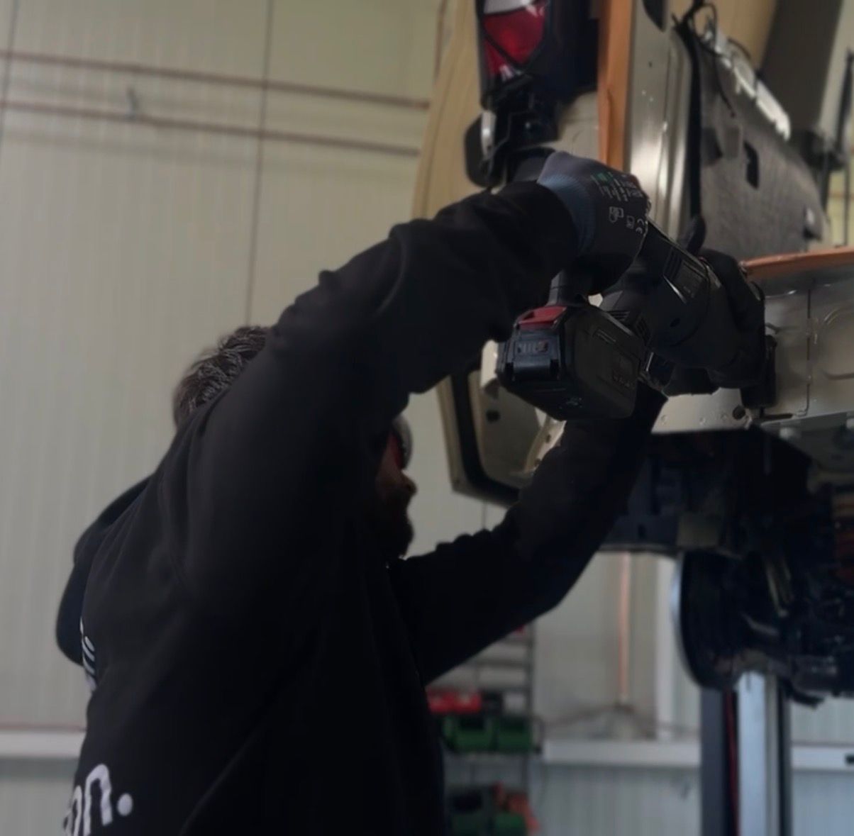 A person wearing gloves uses a power drill to work on the underside of a raised vehicle in a garage.