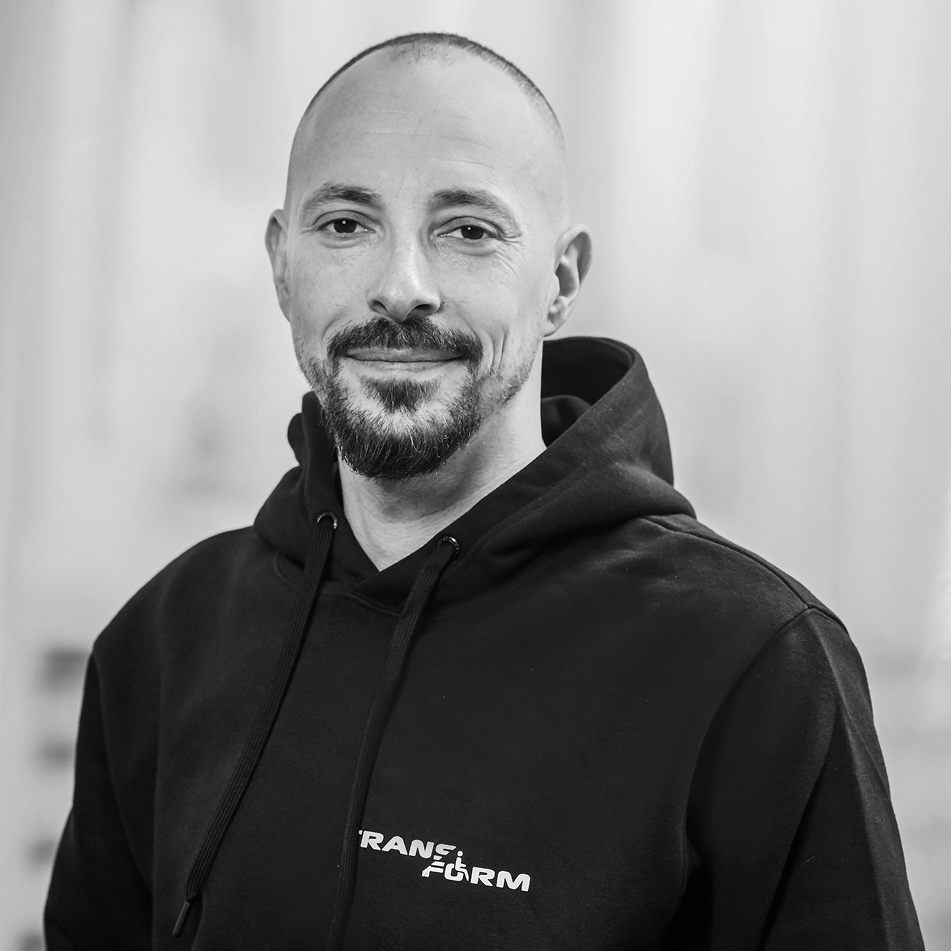 Smiling bald man with a goatee wearing a black hoodie with the word 'TRANSFORM'.