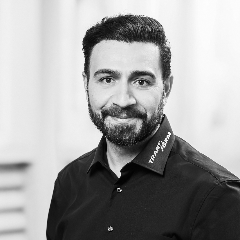 Smiling man with dark hair and beard wearing a black shirt with 'TRANSFIRM' text on collar.