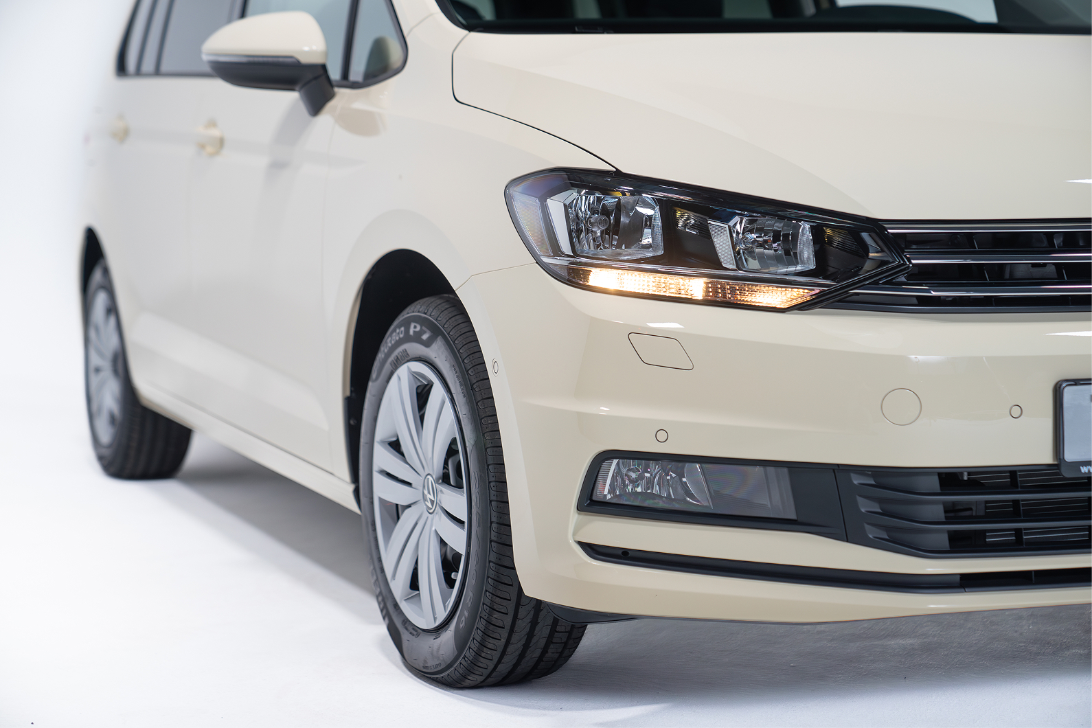 Front-left side of a beige Volkswagen car showing the headlight, grille, and wheel.
