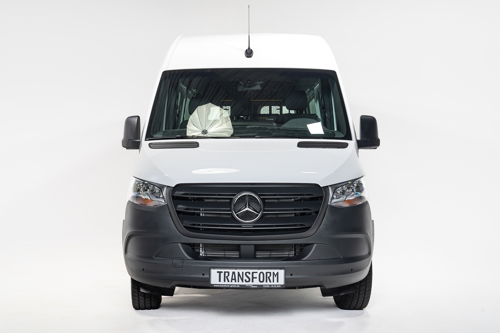 Front view of a white Mercedes van with a black grille and a license plate that reads TRANSFORM.