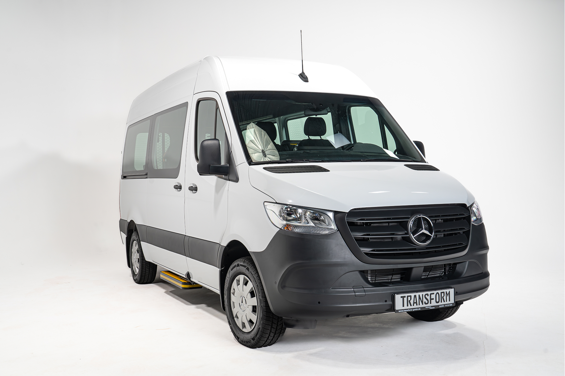 White Mercedes-Benz van with black trim and a front license plate reading TRANSFORM in a white studio.