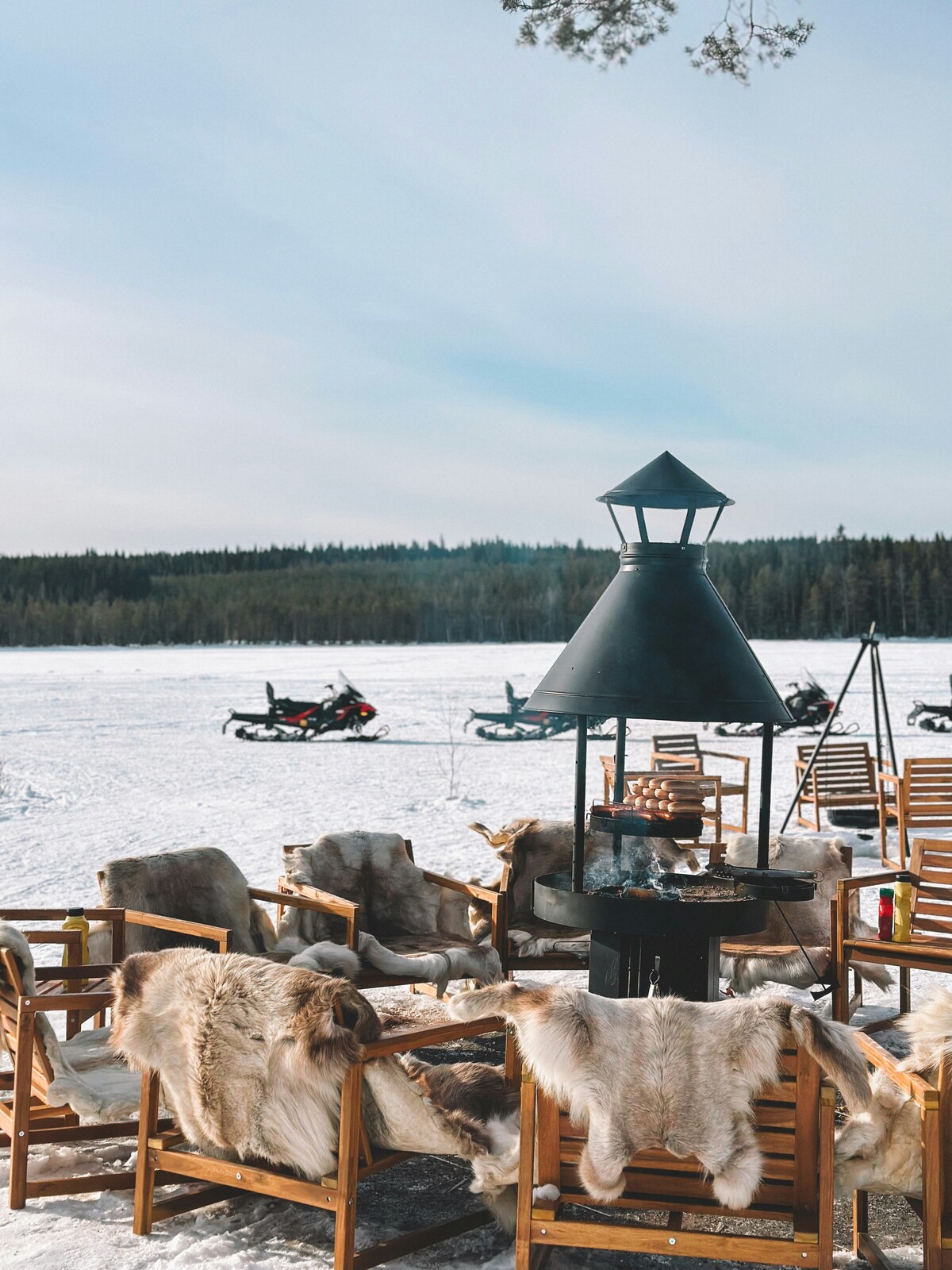 Reindeer-hide chairs around a fire grill by a frozen lake with snowmobiles