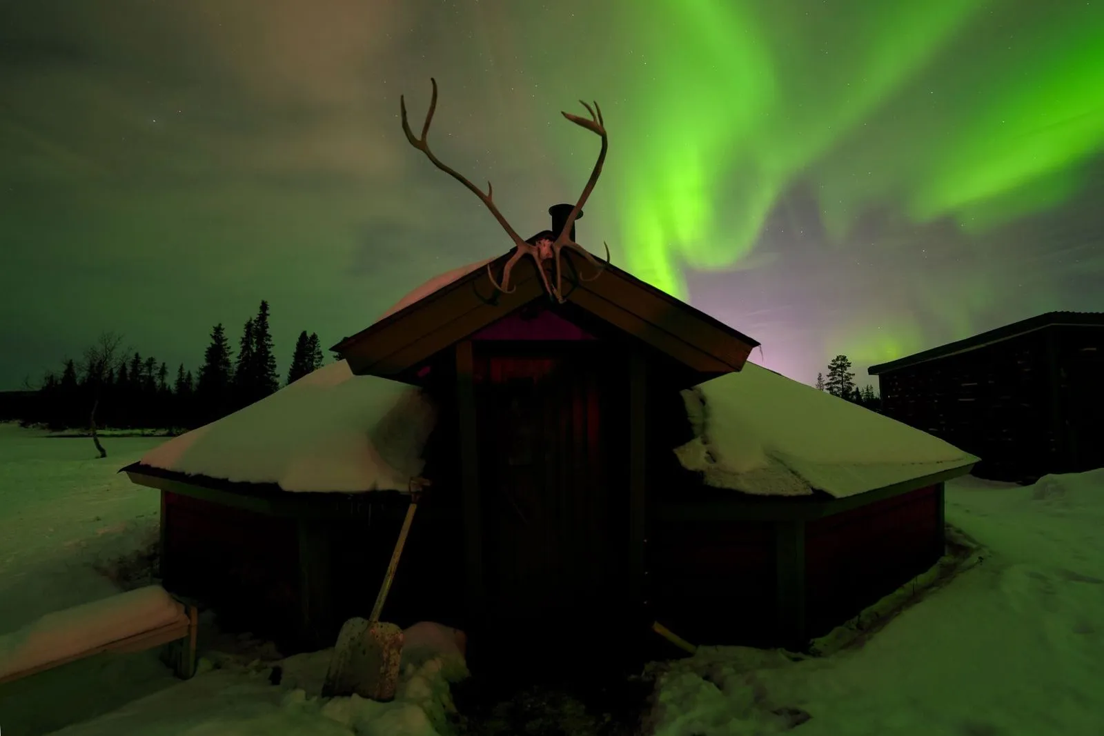 Northern lights glowing green above a snow-covered wooden cabin with reindeer antlers on the roof