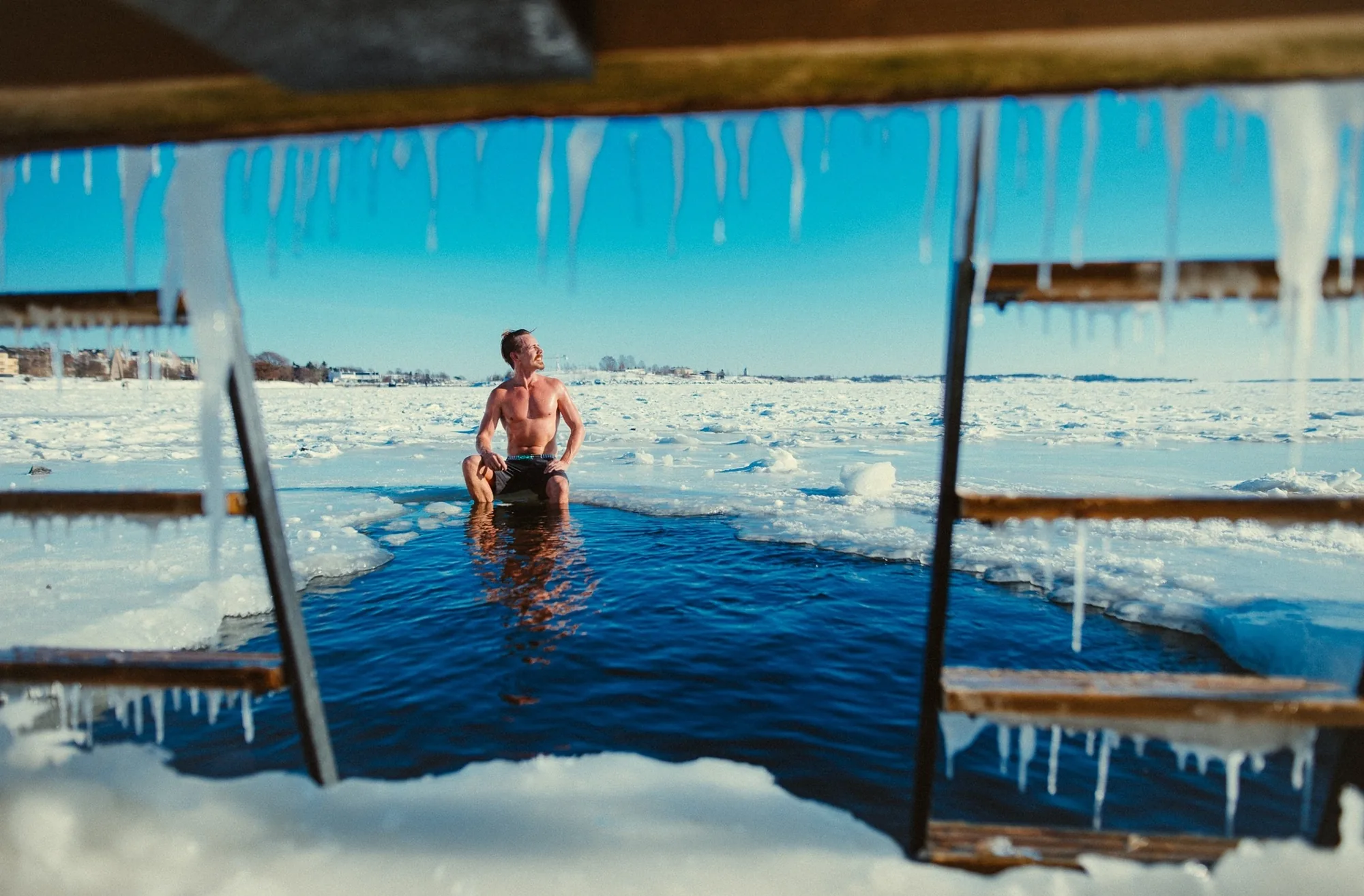 Man sitting in an ice hole in the frozen sea at Löyly in Helsinki, framed by icicles and wooden ladders