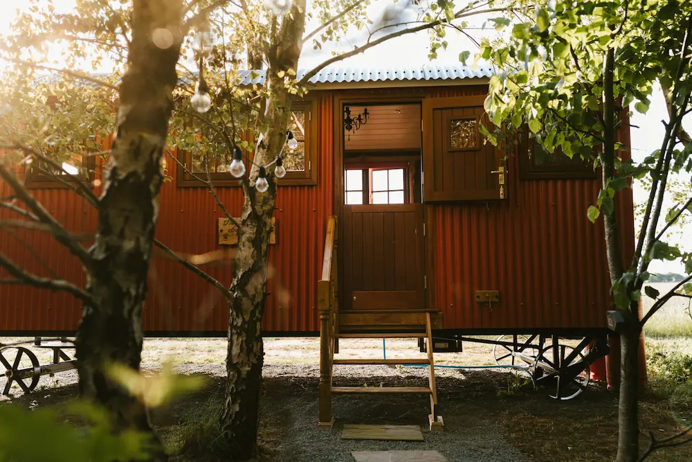 The Badger Sett, a red wooden cabin on wheels with open door, small stairs, trees, and hanging light bulbs in sunlight.