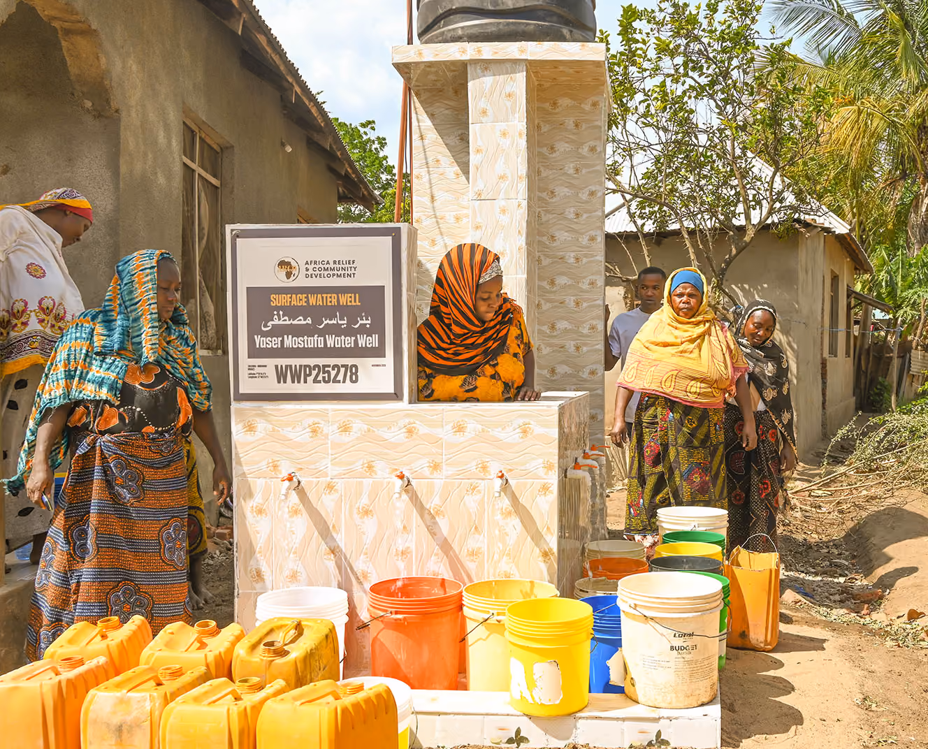 Several people gather around a tiled surface water well with colorful buckets and yellow jerrycans.