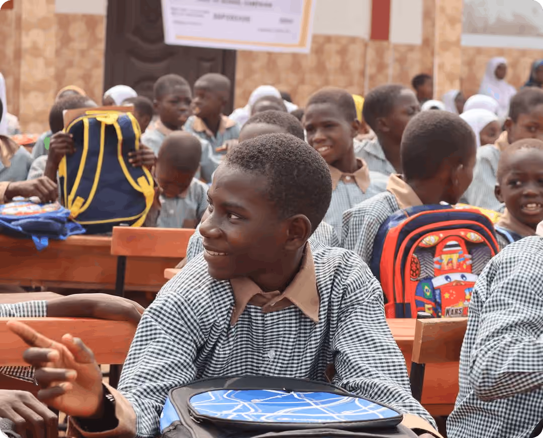 A young schoolboy wearing a patterned uniform shirt sitting at a desk in a classroom with fellow students.