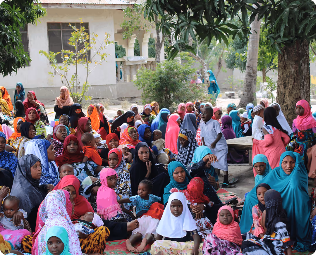 A large group of women and children, many wearing colorful headscarves, seated together outdoors in a shaded courtyard during a community gathering.