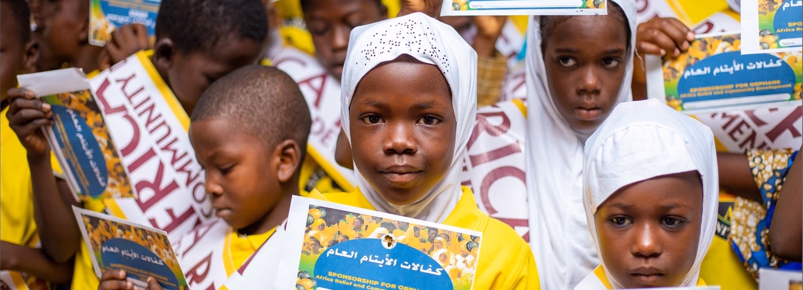 A group of local children and community members posing outdoors in front of an Africa Relief project signboard.